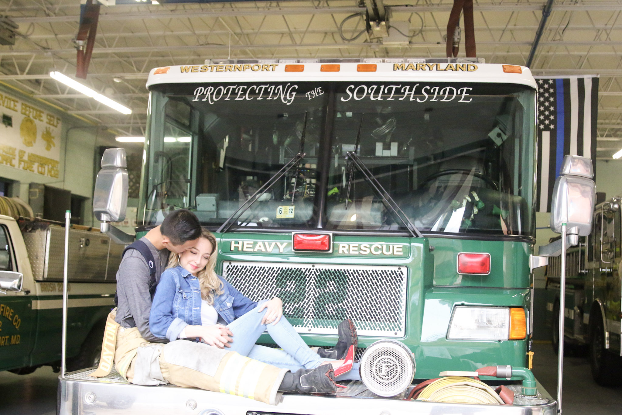 The couple is sitting on the fire truck's bumper. The woman is dressed in eye-catching red high heels, jeans, and a denim jacket, while the man is dressed in his firefighting clothing. 