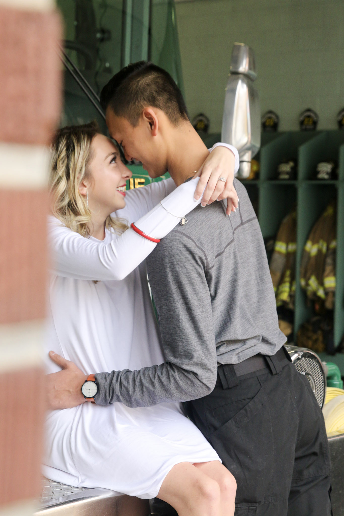 the soon-to-be bride is dressed in a white long-sleeved flowing dress, with her arms wrapped around his neck, while the young man is dressed in a gray long-sleeved shirt and black pants.