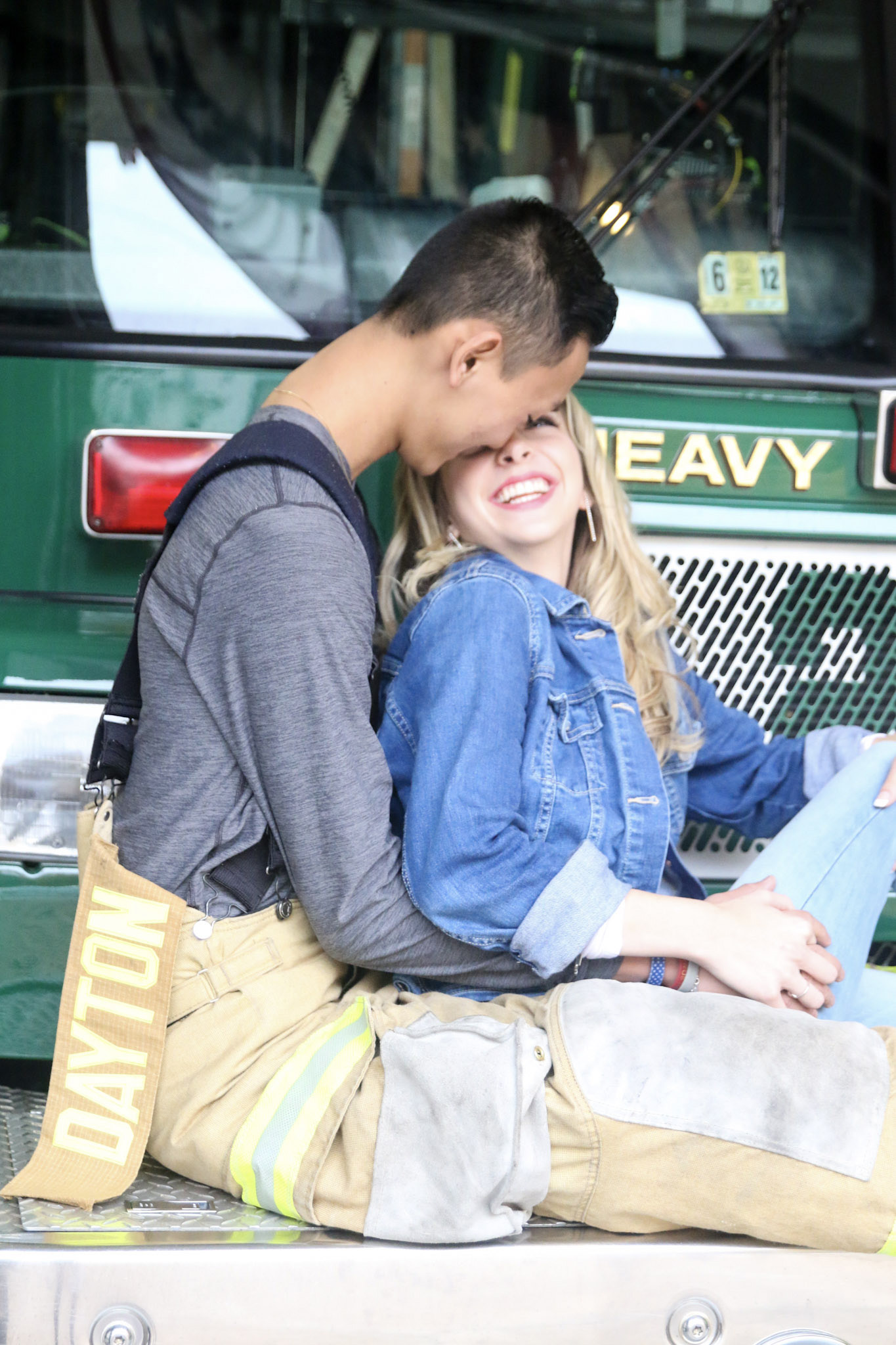 The lady and the man sitting in the front bumper of the fire truck are beaming as the man hugs her from behind.