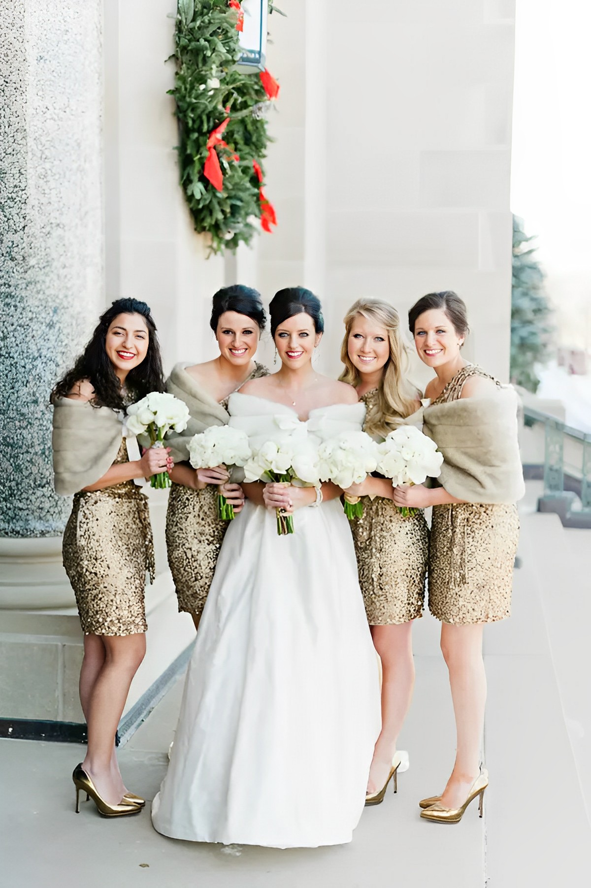 the bride is dressed in a lengthy white wedding gown, while the bridesmaids are wearing shimmery knee-length dresses and holding bouquets along with shawls.