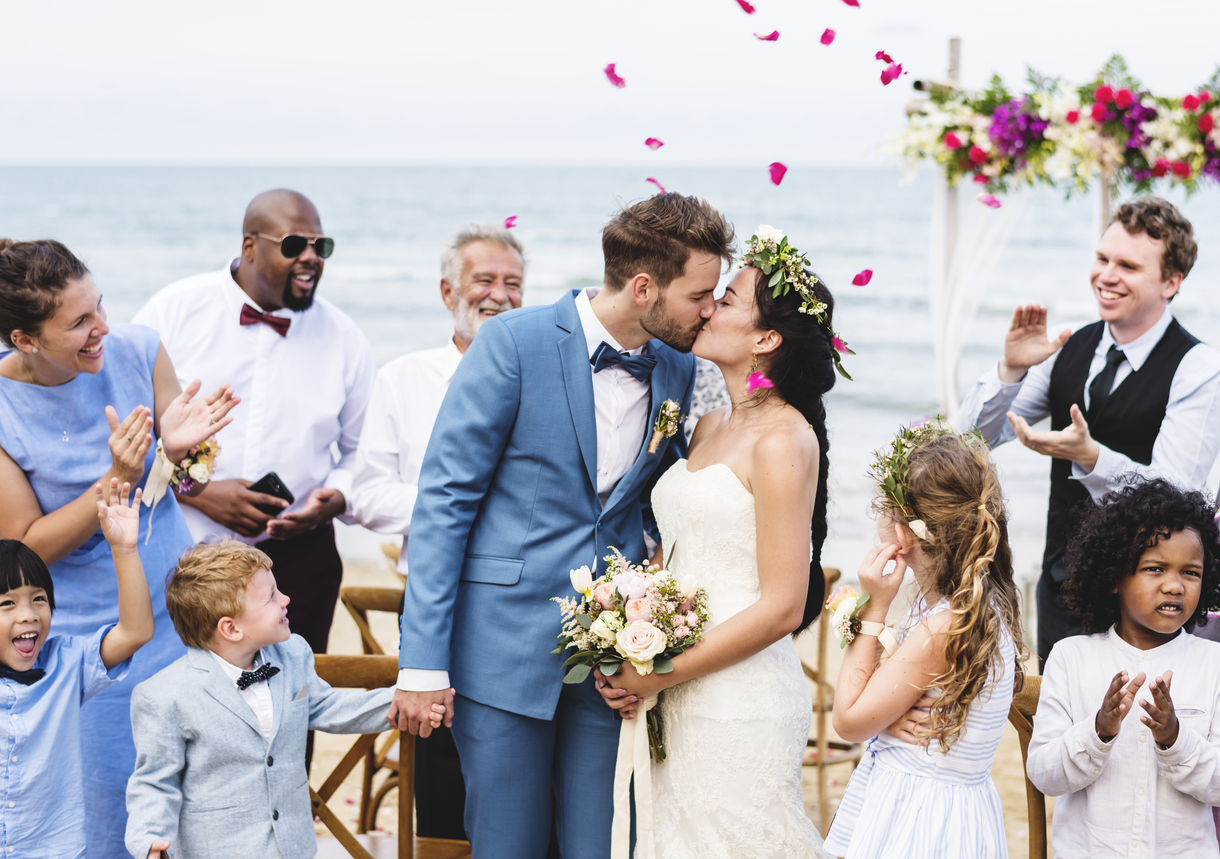 Young couple kissing during seaside wedding reception as friends applaud them on