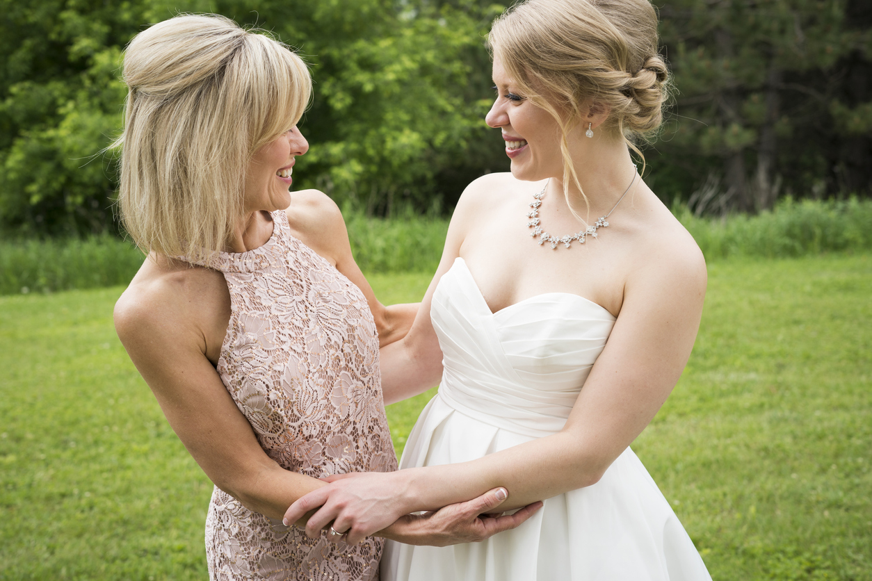 A smiling blond bride in a white wedding gown and her lovely mother in law in a pink pastel lace gown enjoy a tender moment at an outdoor wedding