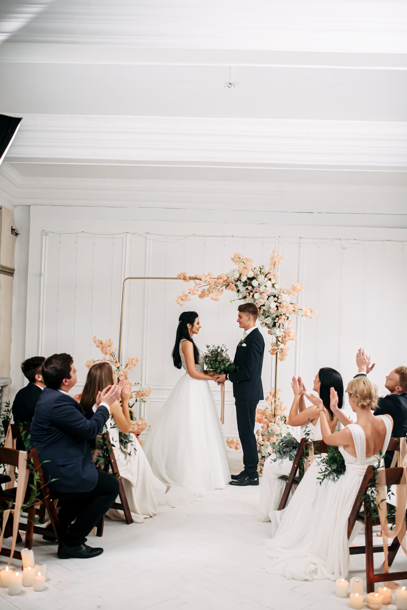 Indoor boho style wedding arch. A couple of newlyweds in front of the altar hold hands and look at each other with love. Guests are sitting on chairs