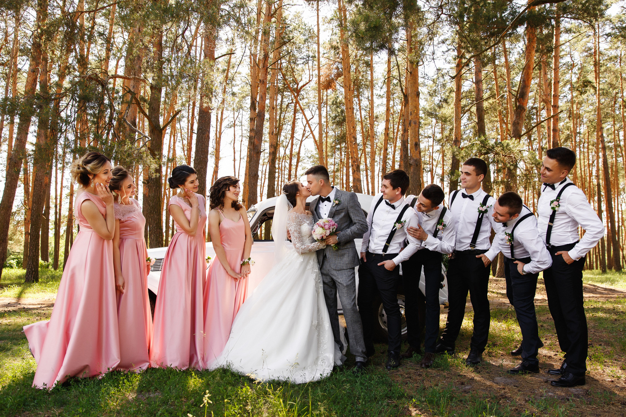 Group wedding photo. Bride and groom kisses near friends, bridesmaids in pink dresses and groomsmen with bow ties and suspender looks at newlyweds