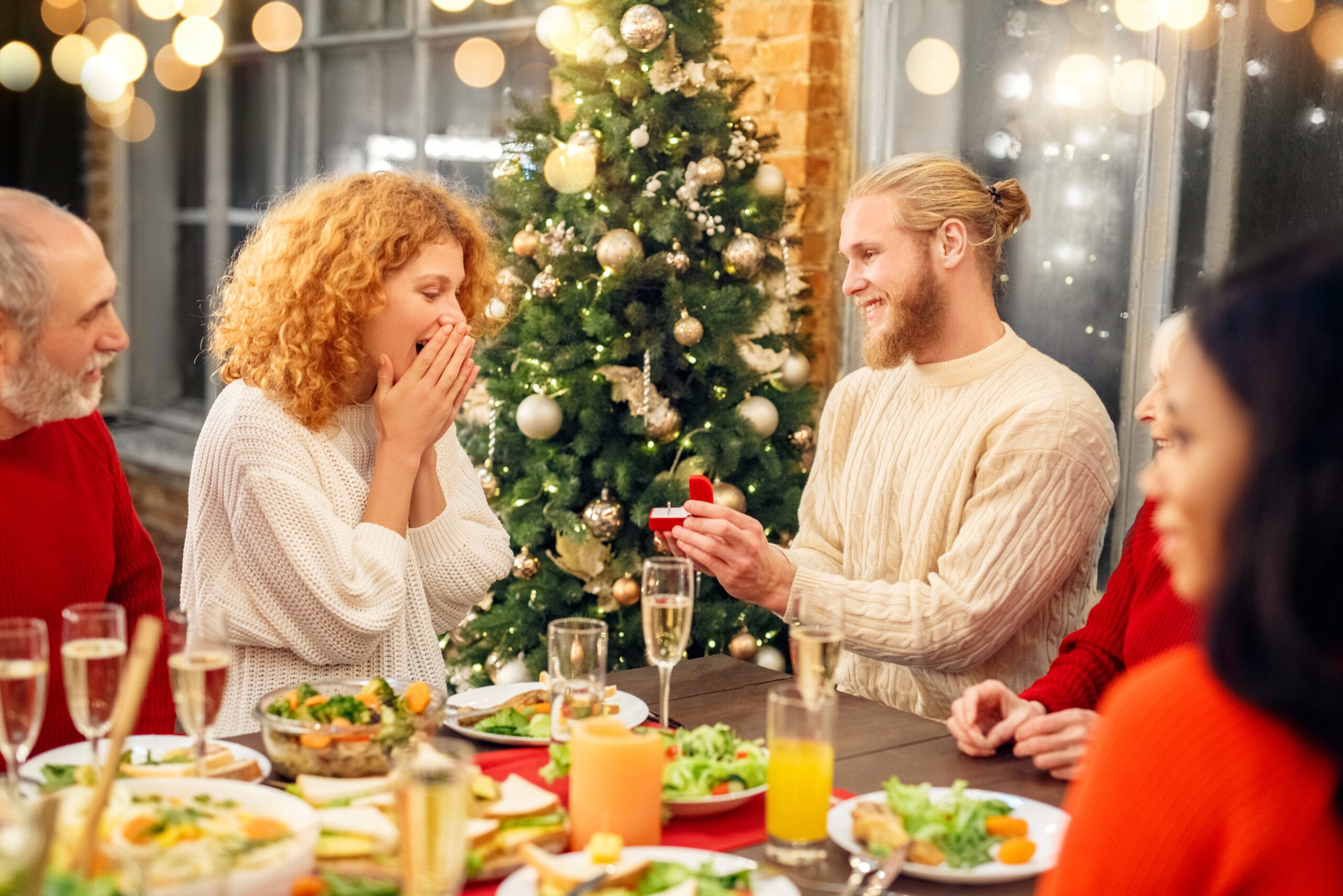 Smiling young man making proposal with ring in box to happy surprised curly woman at multiracial family Christmas dinner with relatives at festive table over home and Christmas tree background