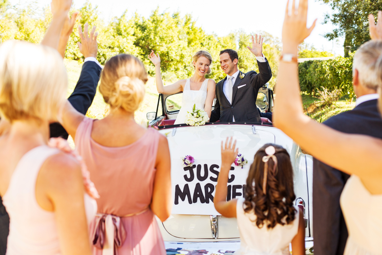 a happy newlywed couple in a convertible waves to wedding guests while holding a placard that reads "Just Married."