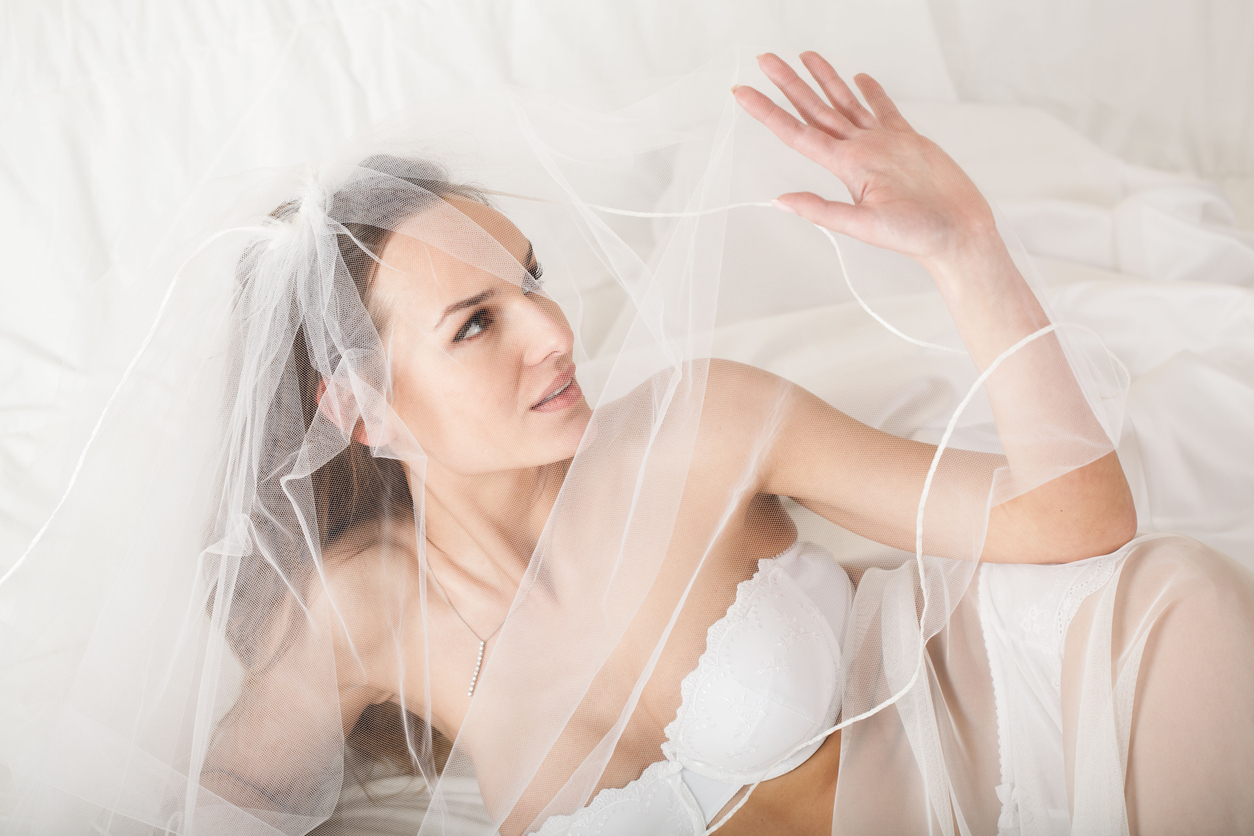 Young woman in bridal lingerie lying in bed and covering her face using veil