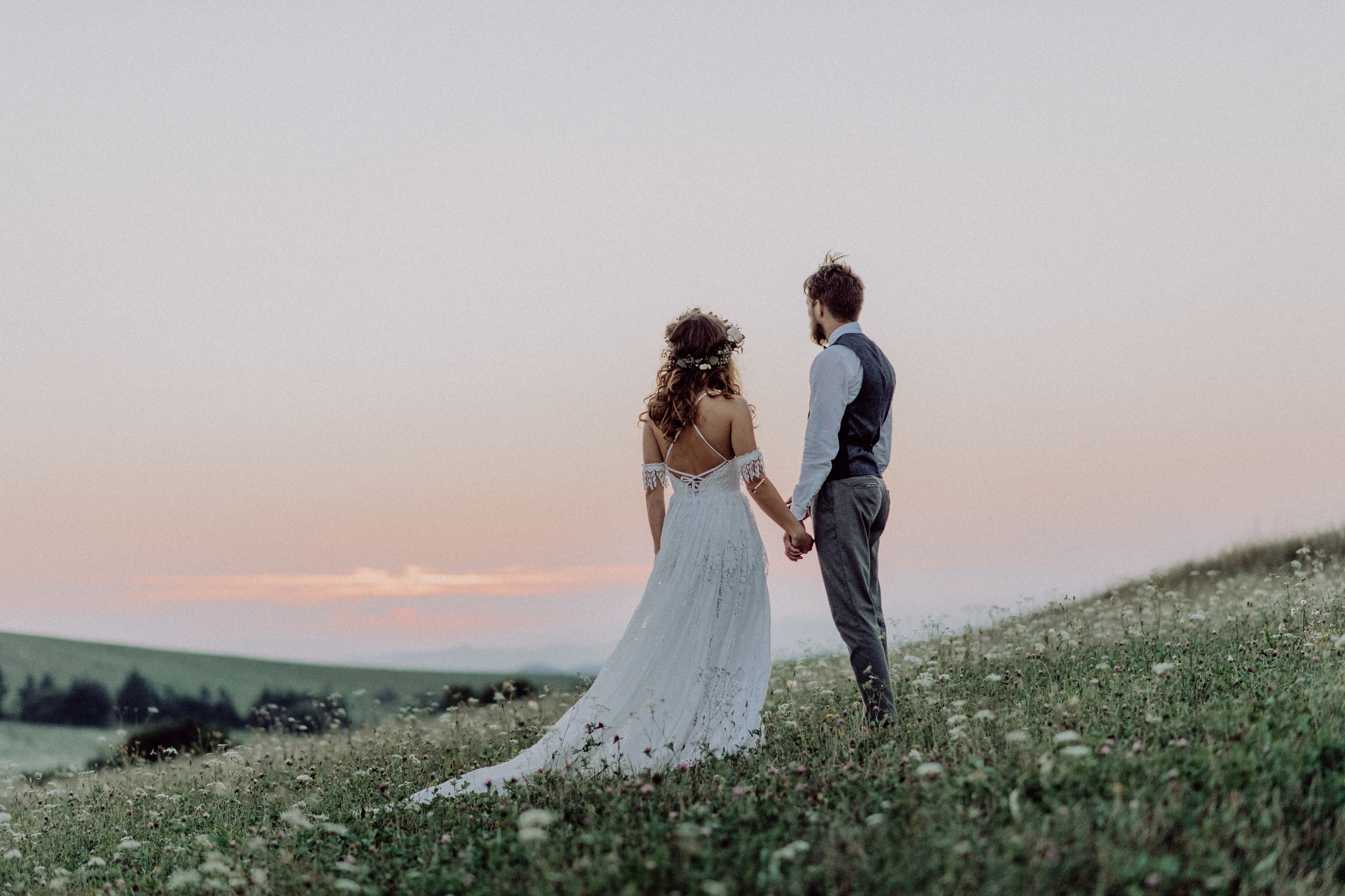 Beautiful young bride and groom outside in green nature at romantic sunset, holding hands. Rear view.