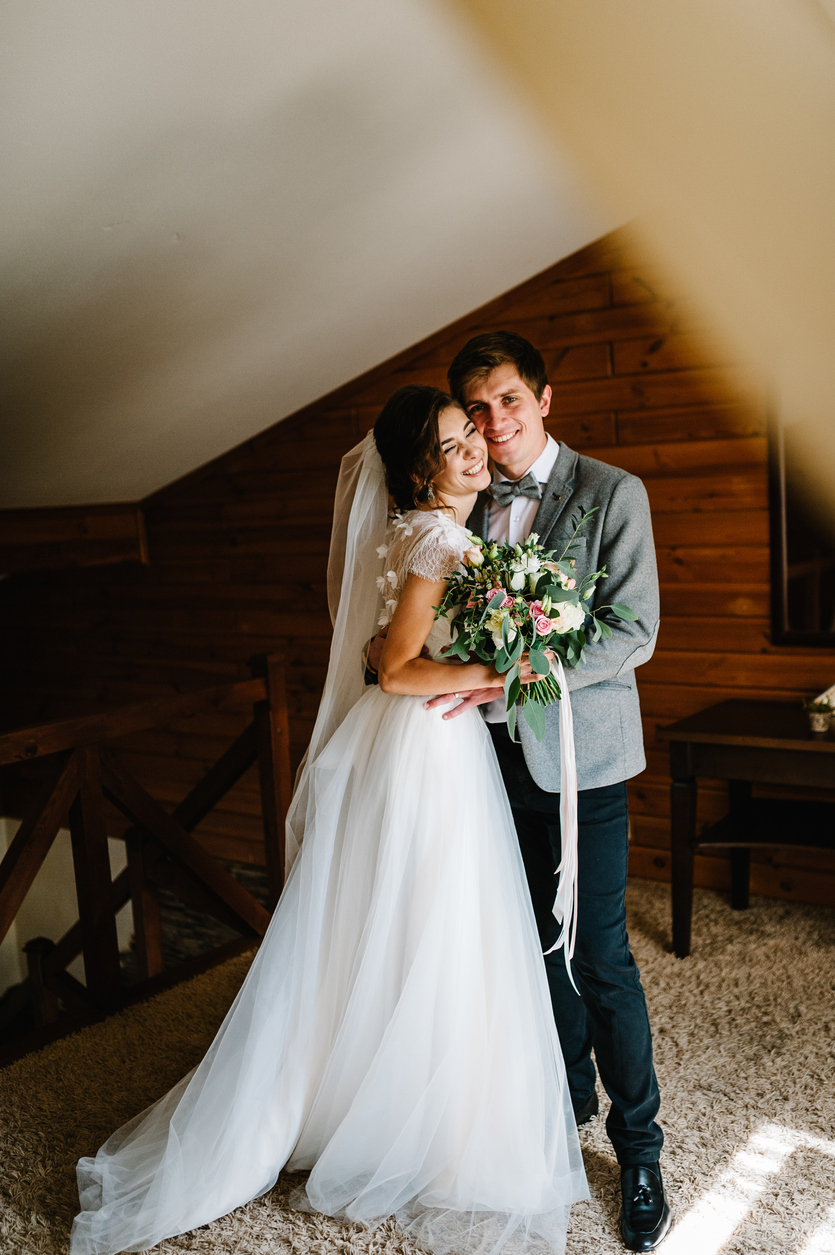 The groom in suit hugs bride in white dress are holding a beautiful wedding bouquet flowers, greenery and decorated with ribbon at home. Portrait of a beautiful young couple.