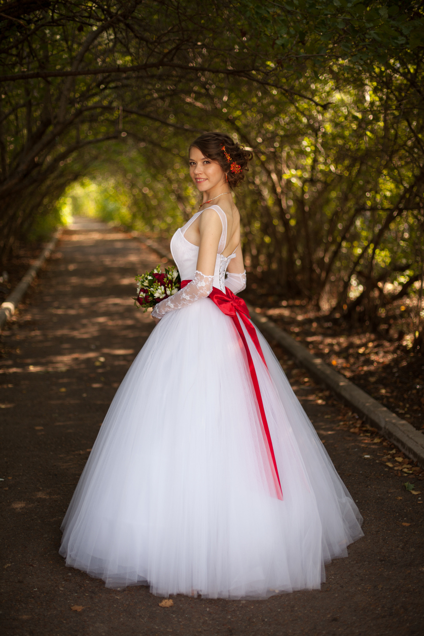 bride in an outdoor shoot with her long white wedding dress accentuated with a red ribbon and her bridal bouquet.