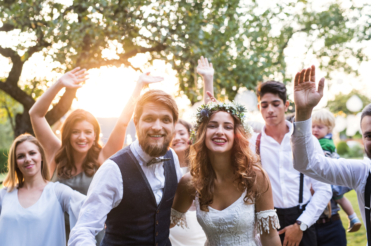 The bride and groom are in the middle of the photograph, with their wedding guests beside and behind them, posing for a sunset photograph at a wedding reception.