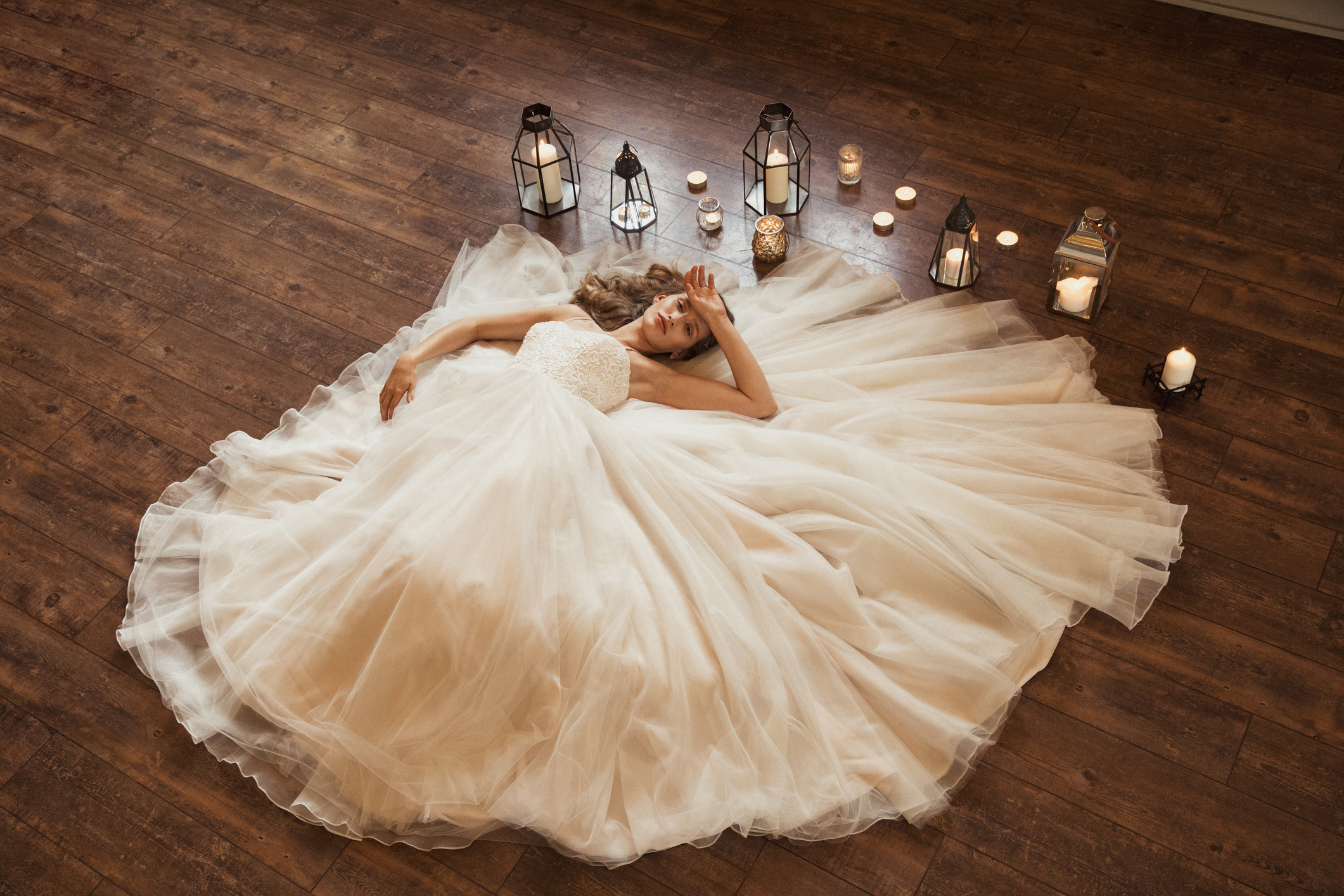 Bride laying on a wooden floor in her wedding dress with one hand on her head. There are different candles spread out around her.