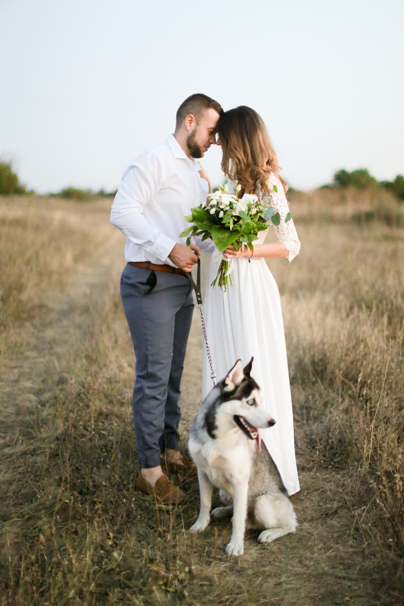 groom hugging bride in white dress with bouquet of flowers near husky in steppe background