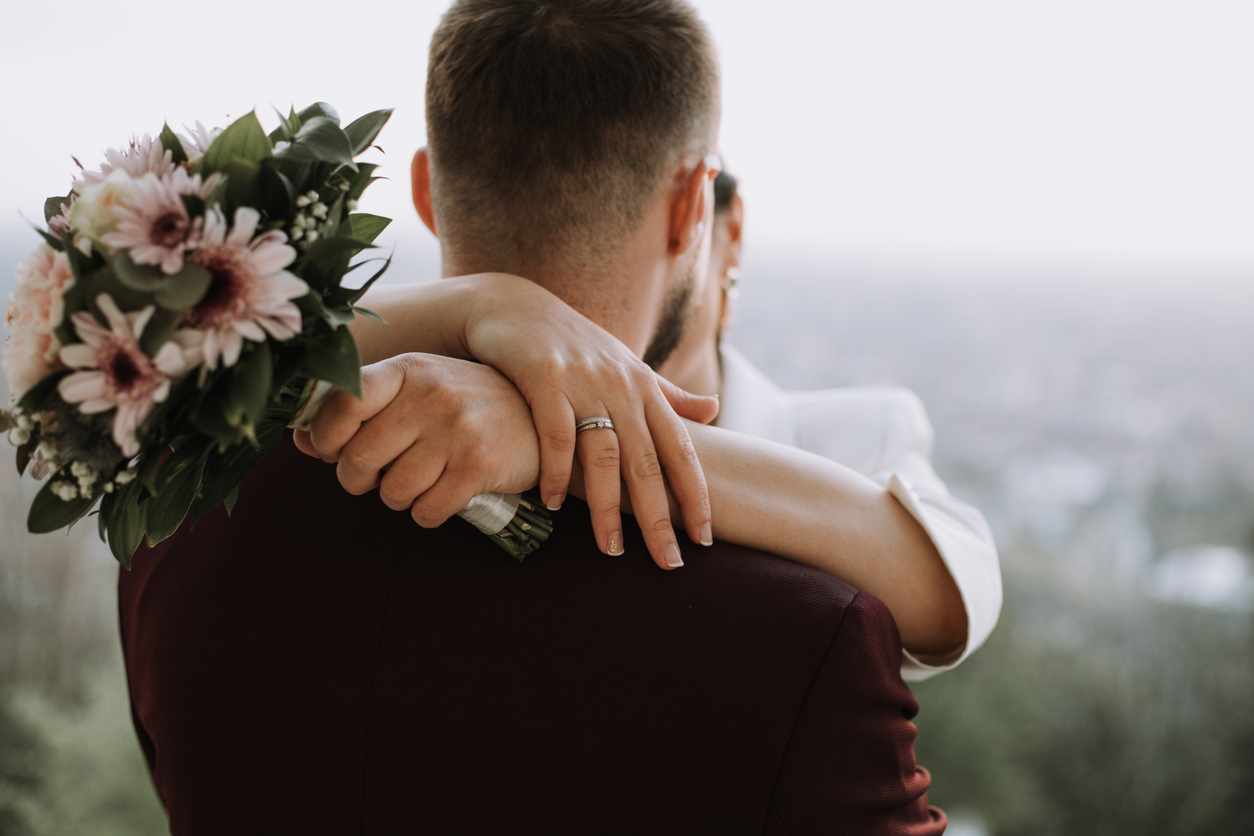 The groom is shown from behind as the bride wraps her arms around him, displaying her ring and wedding bouquet.