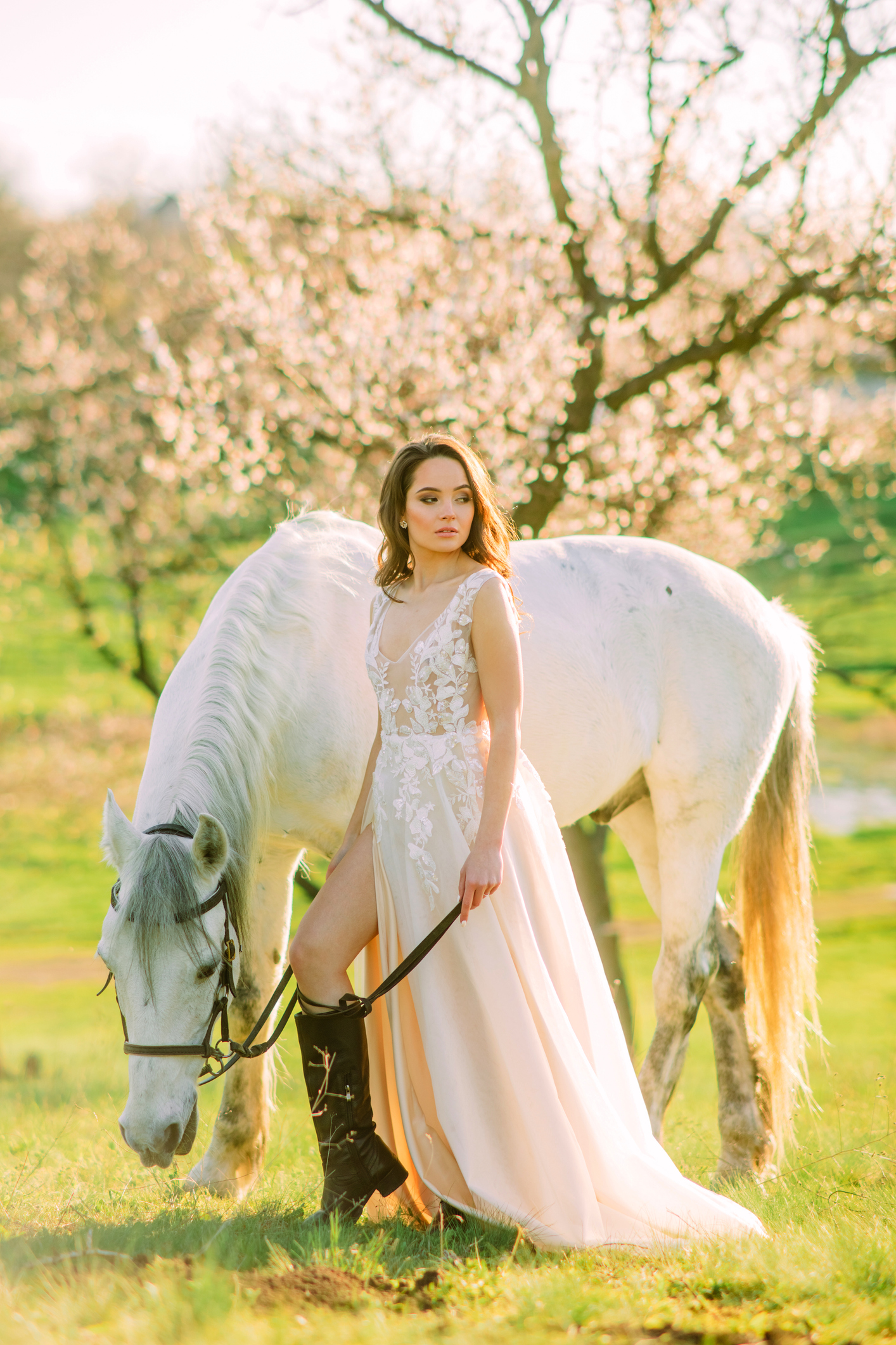 bride in white dress stands near horse in spring blooming garden