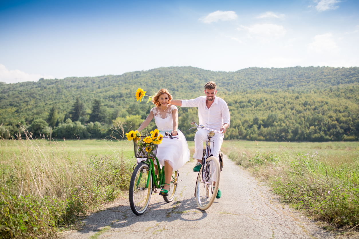 a youthful couple is having a good time riding a bicycle  through a meadow, and the woman has a sunflower placed in the basket of her bicycle.