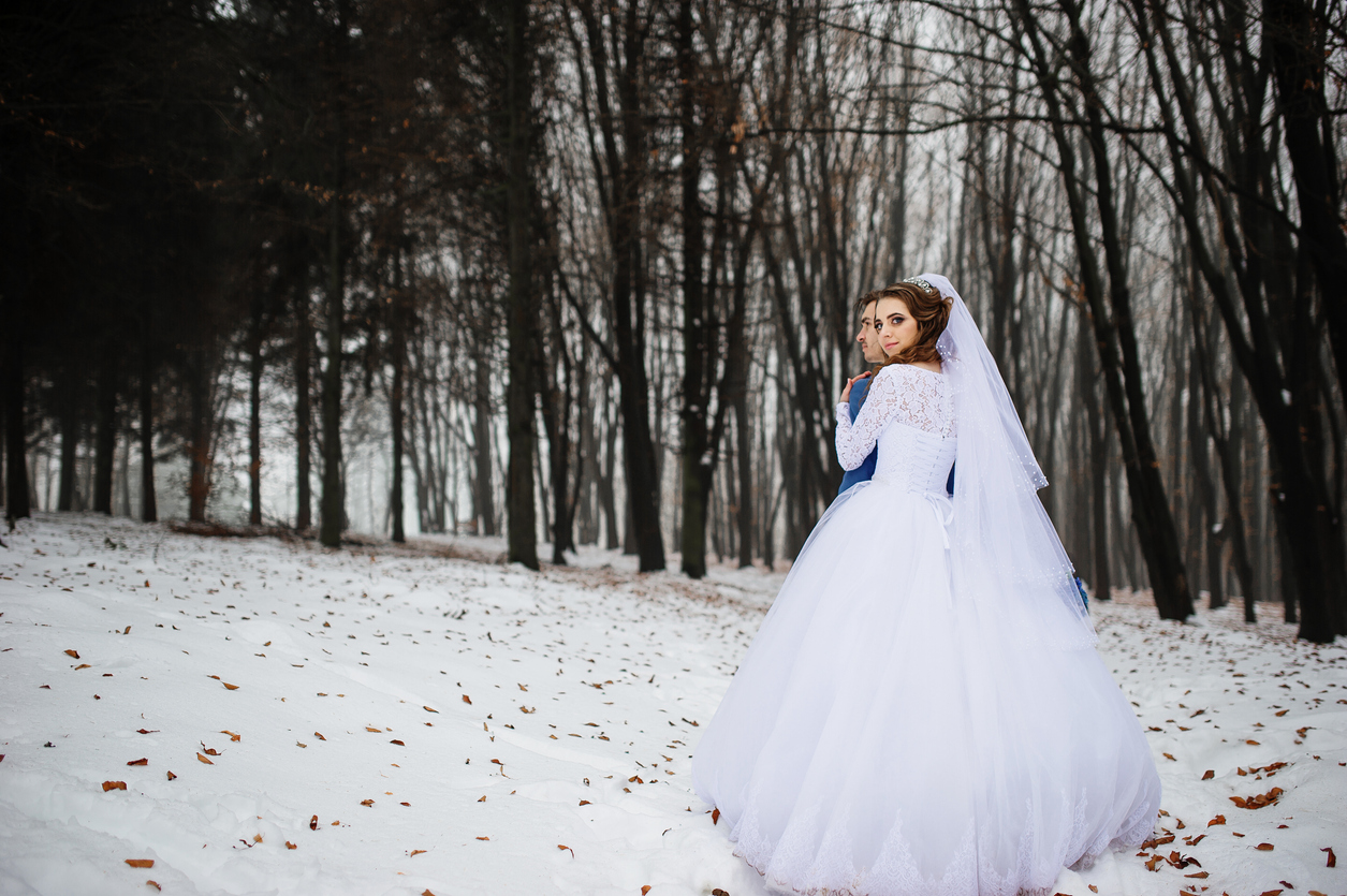 In a snowy field surrounded by trees, the bride in a long-sleeved gown grips the back of her spouse in a blue suit.