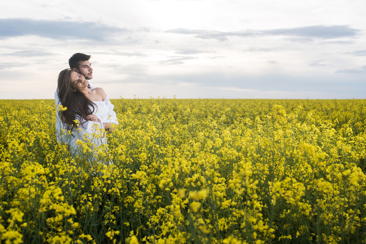 loving couple embracing while standing in a filed full of yellow flowers and looking away. 