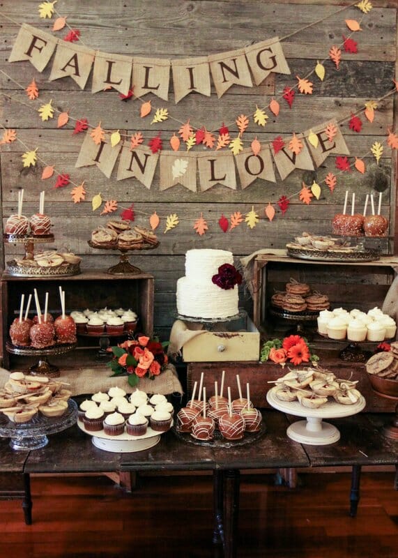 A collection of sweets and desserts are presented on a table, adorned with a banner that says "Falling in Love" and decorated with autumnal green and orange leaves.