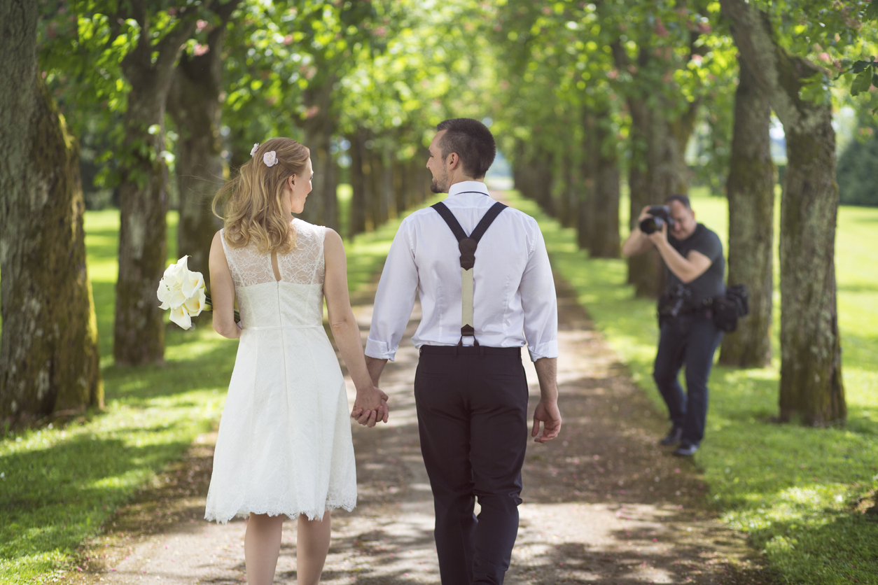 Bride and groom walking in tree-lined path with a photographer in front.