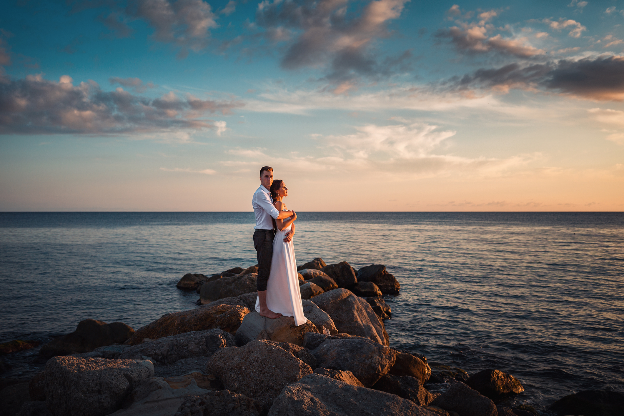 newlyweds embrace and pose standing on the coastal rocks. In the background, the sunset and the ocean and the sky are in the clouds