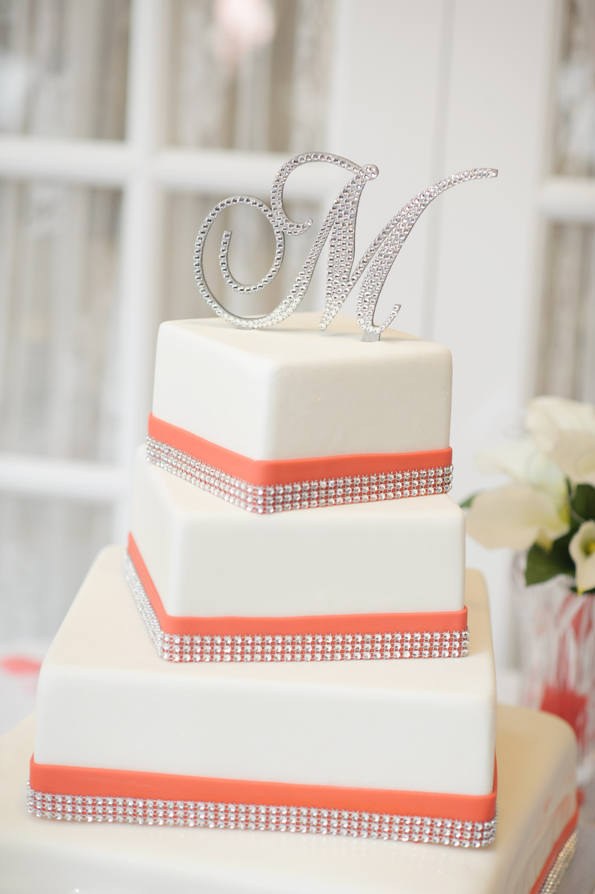 A three-tiered wedding cake with a simple design, crystal decorative elements, and a letter M topping.