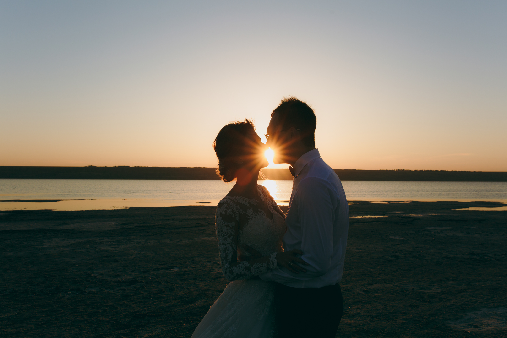 Beautiful wedding photosession. Handsome unshaved groom in a black trousers and young cute bride in white lace pattern dress with exquisite hairstyle on walk along the coastline near the sea sunset