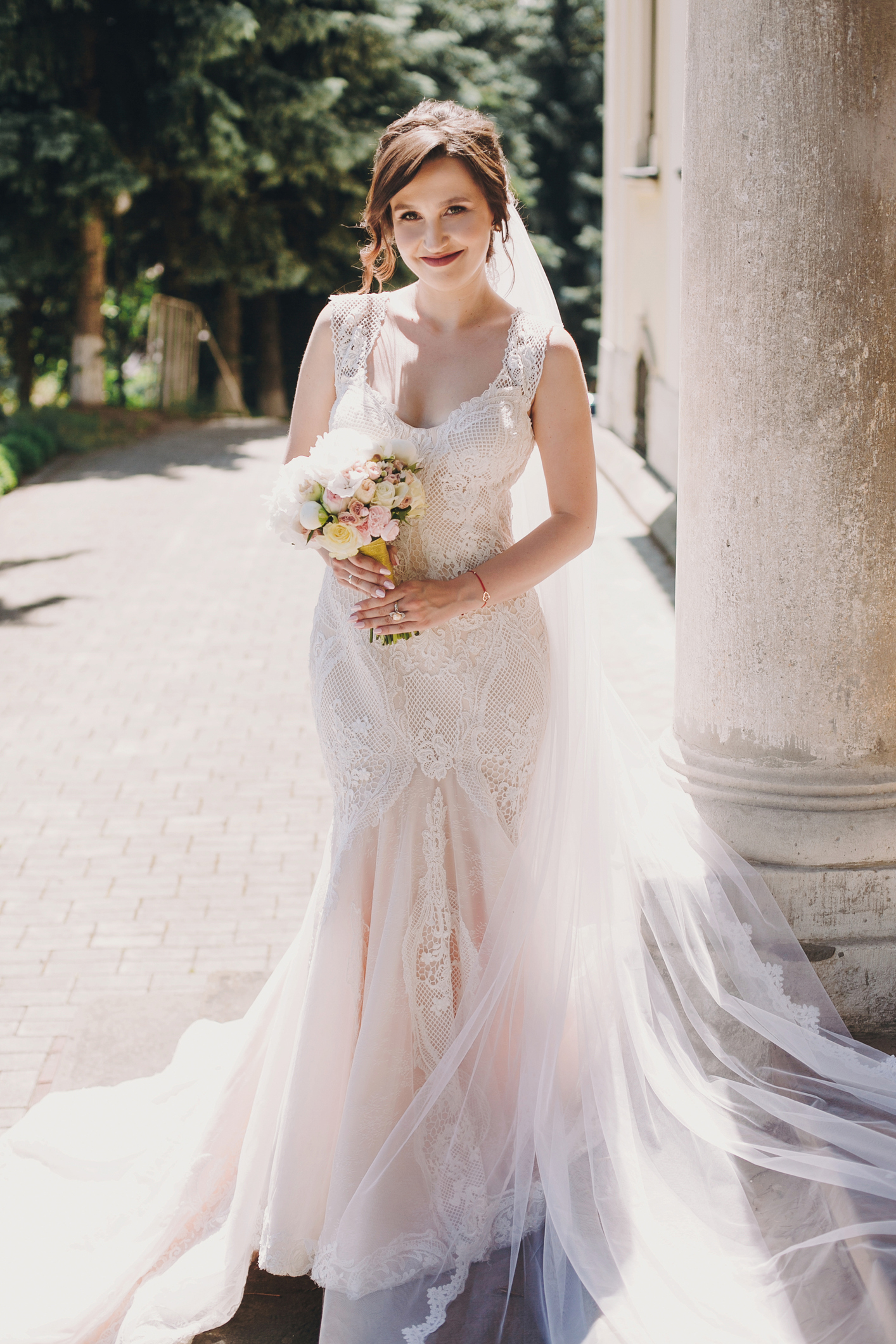 Gorgeous bride in amazing gown with long veil and loop, holding wedding bouquet and posing in sunny city street at old building. Beautiful happy woman in luxury gown.