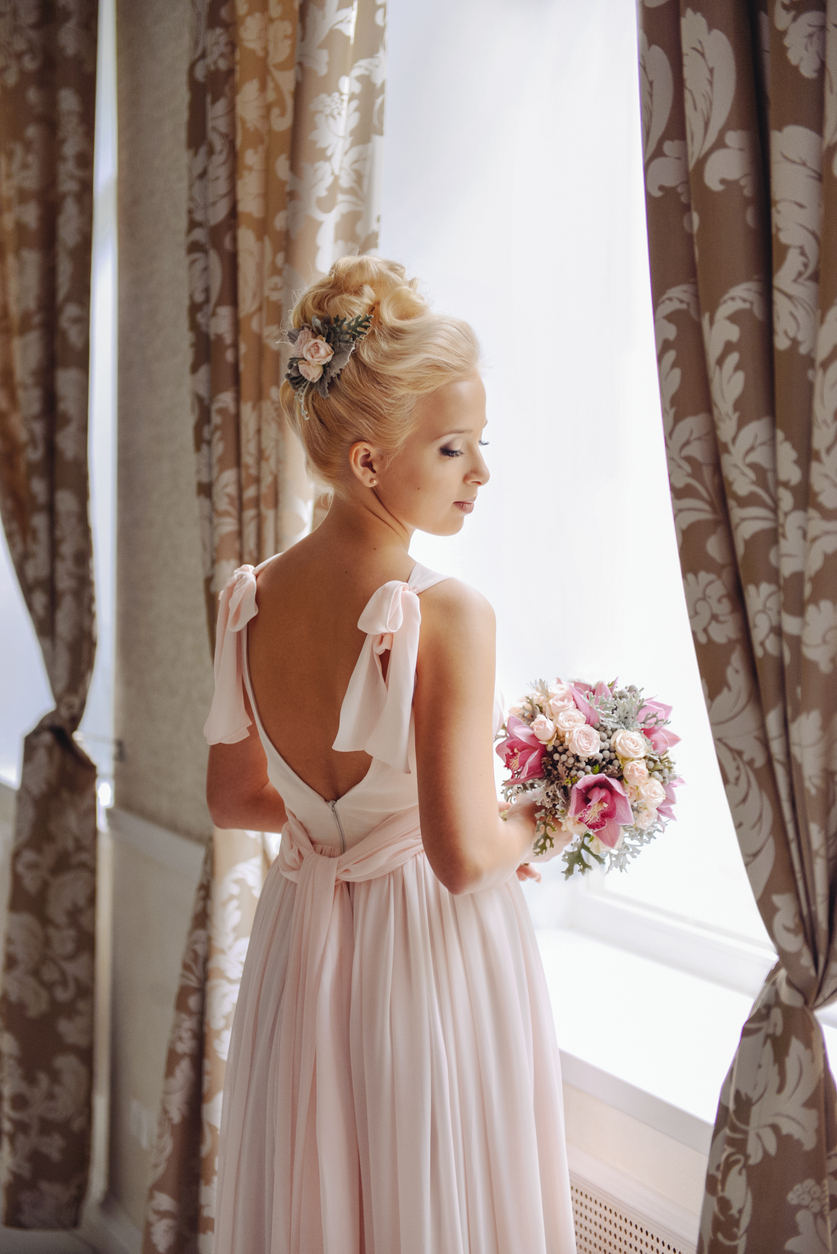 Beautiful blonde young bride standing near the window in a dress with a bare back, holding a bridal bouquet of roses and orchids and a gorgeous floral accessory in her head.