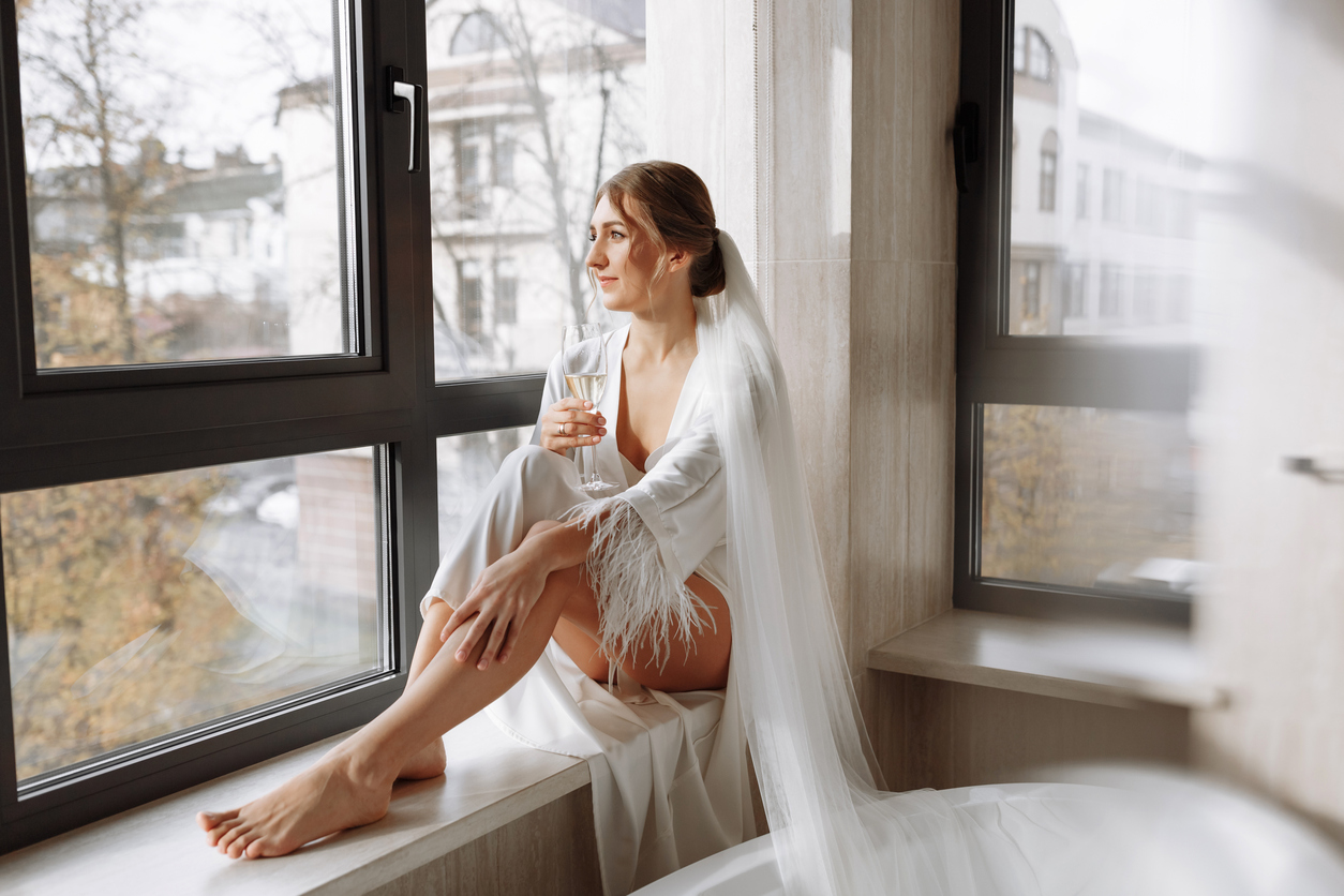 In the peignoir, the gorgeous bride is drinking champagne sitting on a wide window in the bathroom of a hotel room.