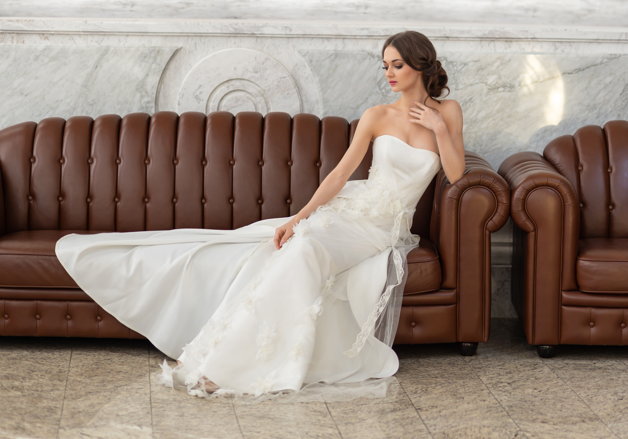 Young fragile brunette. The bride in a luxurious wedding dress is sitting on the sofa in beautiful sunlight from the window. Wedding hair and makeup. Photo for a beauty salon