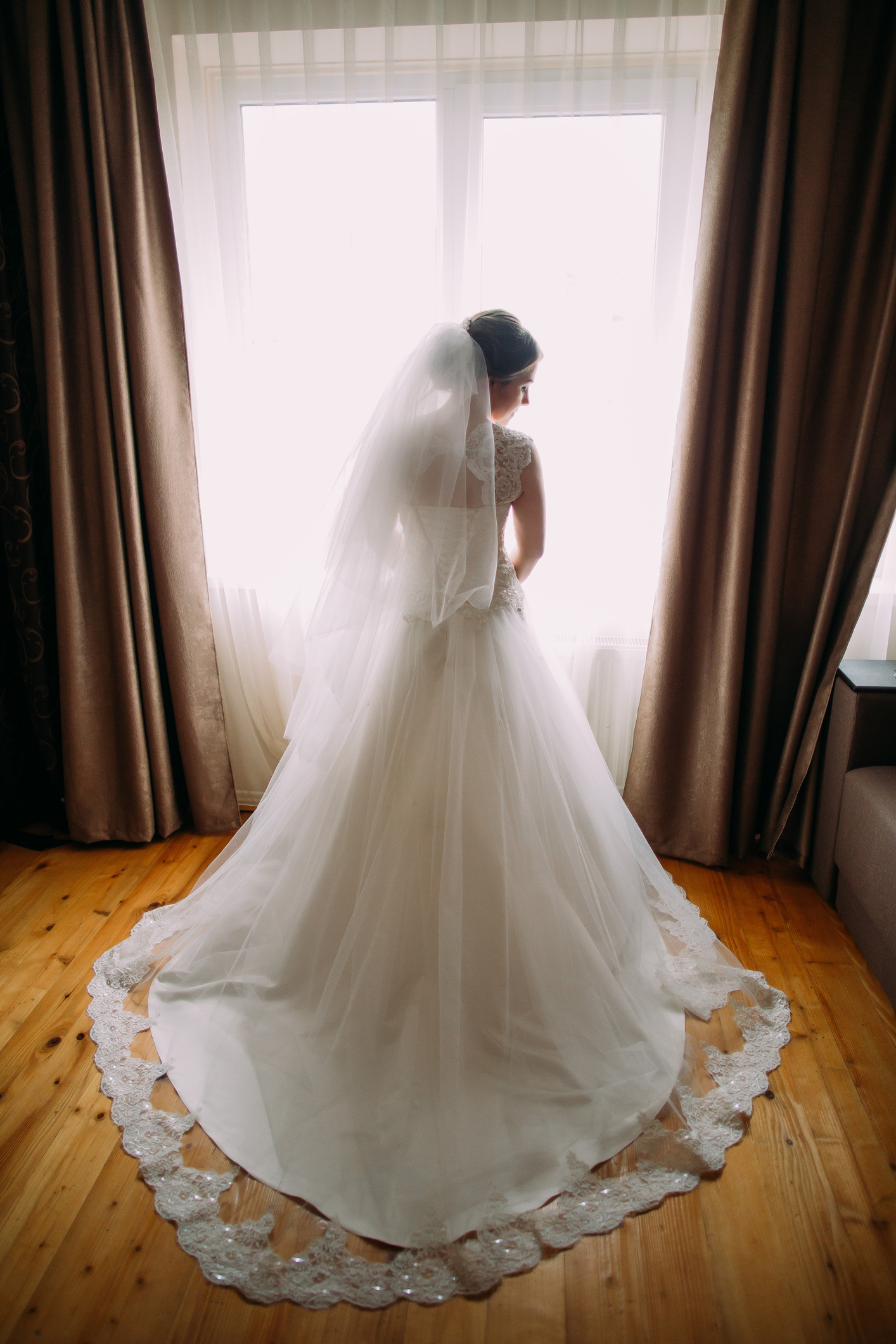 beautiful bride with a long veil wearing a classic wedding gown, standing against window lights