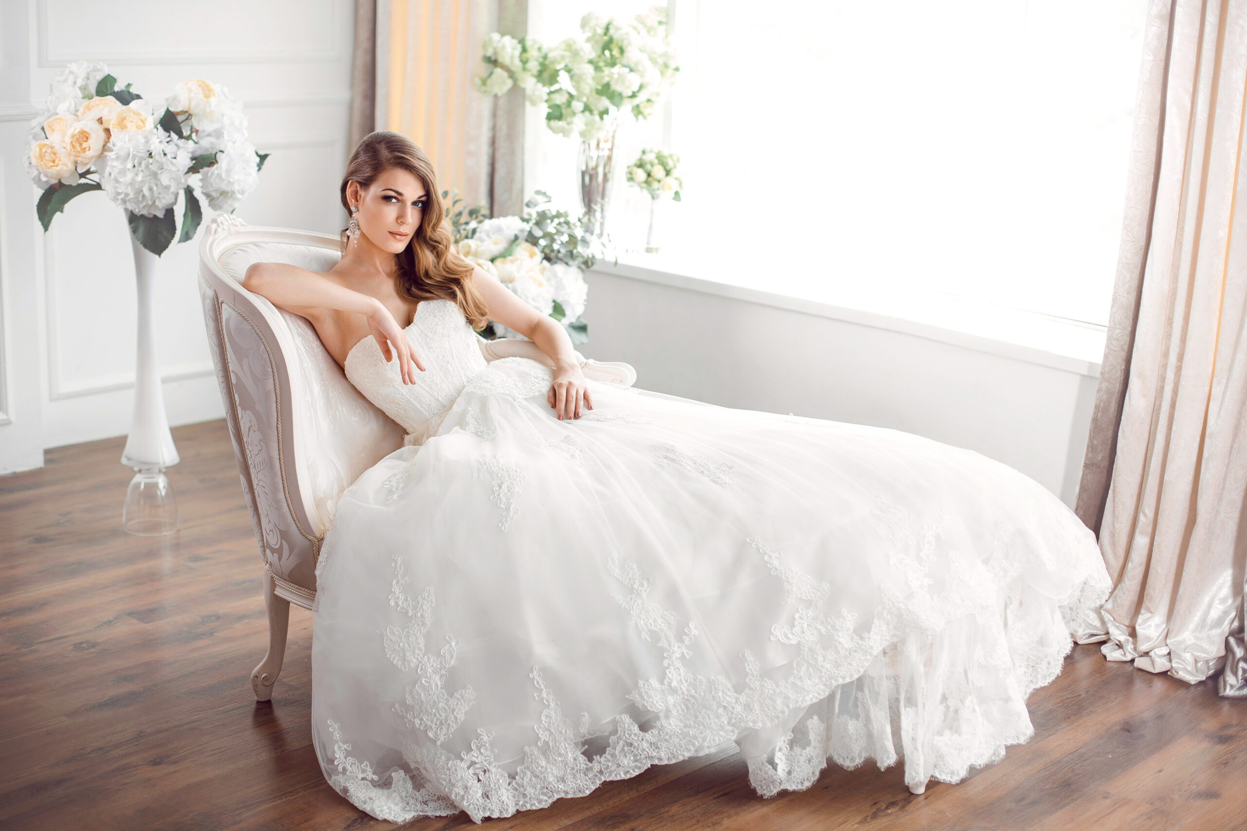 Wedding. Bride in beautiful dress sitting on sofa indoors in white studio interior like at home. Trendy wedding style shot in full length. Young attractive caucasian brunette model like a bride against big window tender posing.