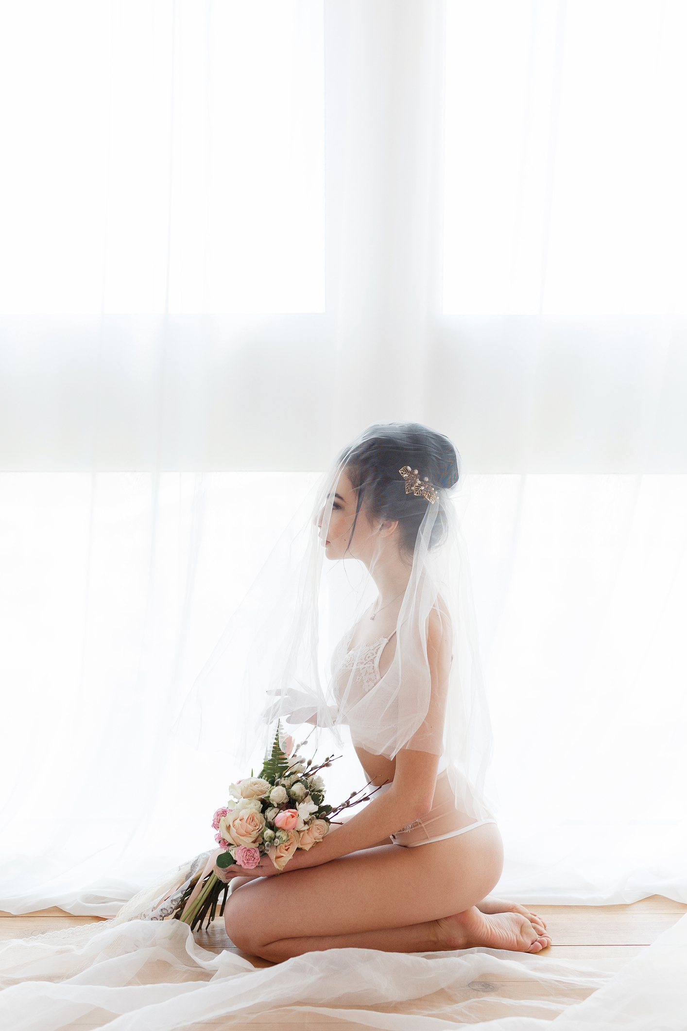 young bride in a white lingerie sitting on the floor next to the big window, covered with veil, morning preparation before wedding