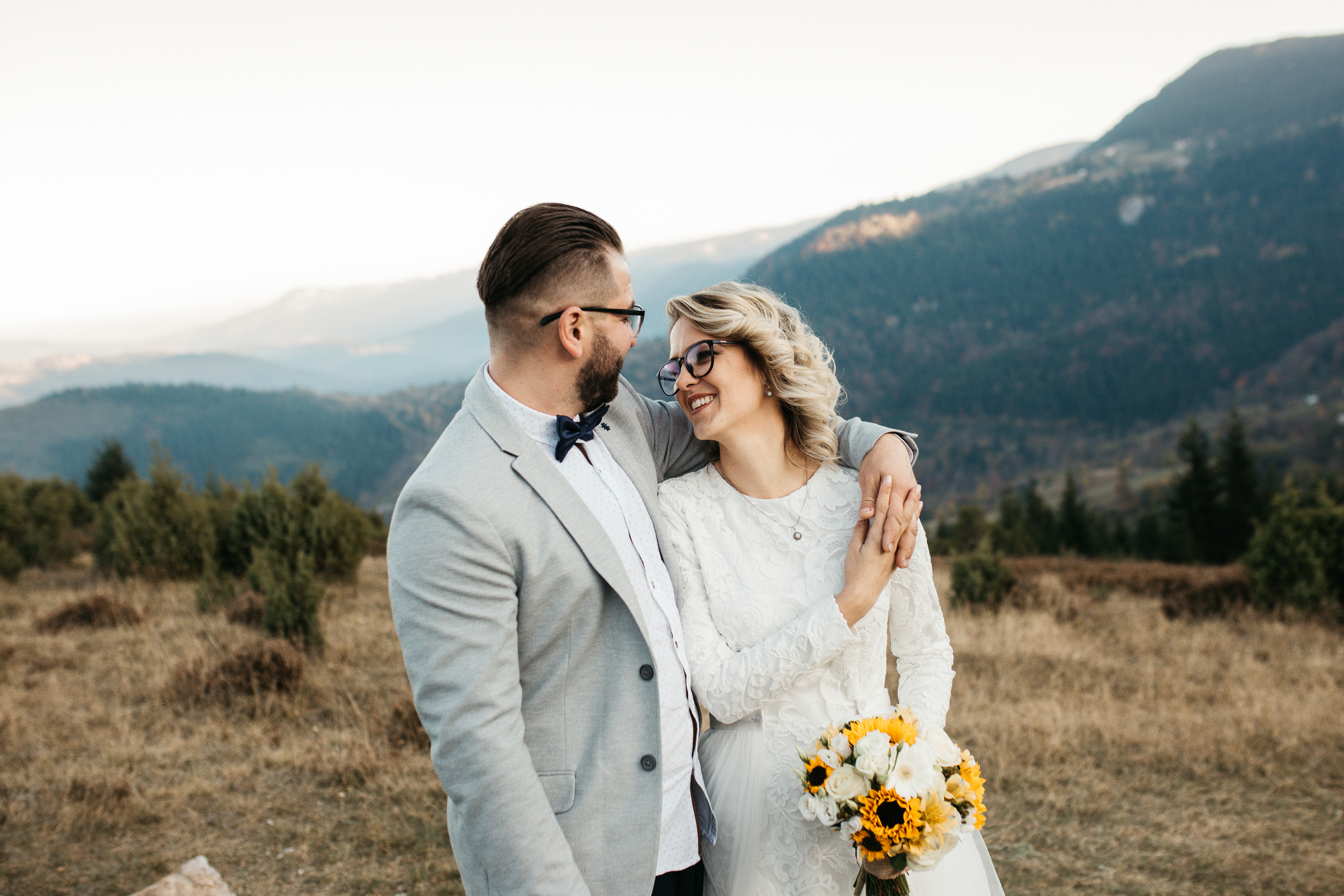 Beautiful couple walking and smiling on their weeding day, in mountains at sunset. Bride is in a white wedding dress with a bouquet of sunflowers in hand, groom in a suit.